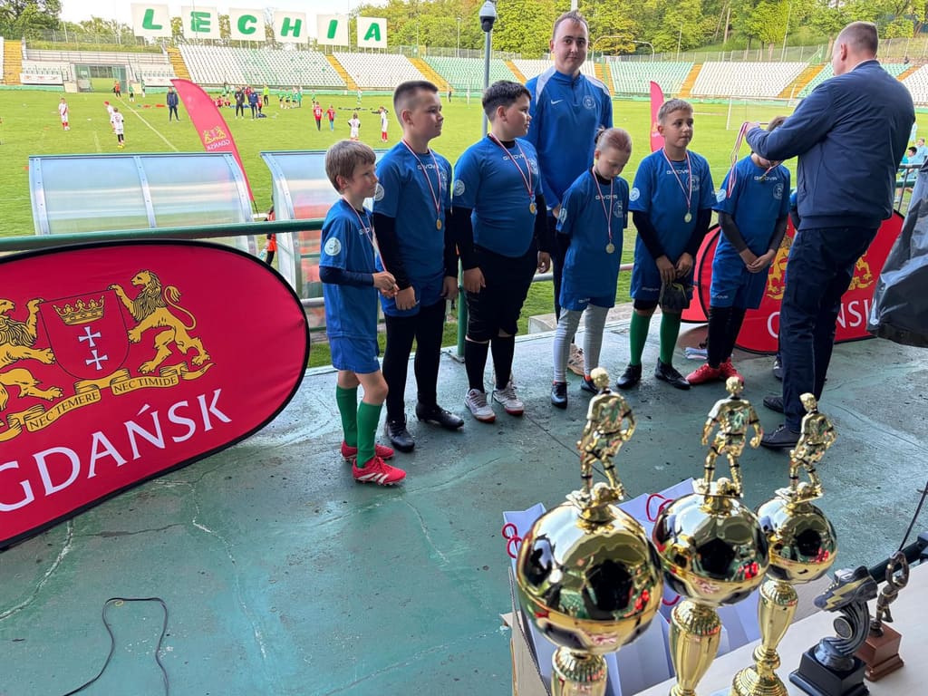 Zawodnicy w niebieskich strojach odbierają wyróżnienia na płycie stadionu Lechii, na pierwszym planie trzy puchary.