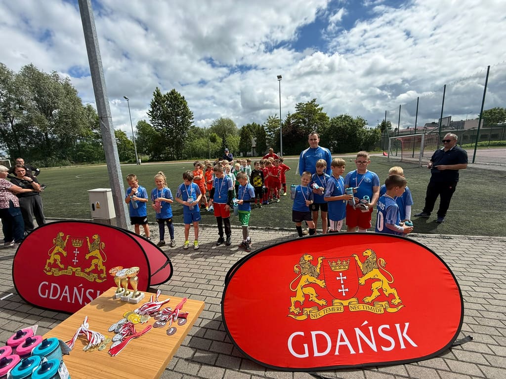 Uczestnicy ustawieni w szeregu przed dwoma czerwonymi banerami z herbem Gdańska i stołem z medalami.
