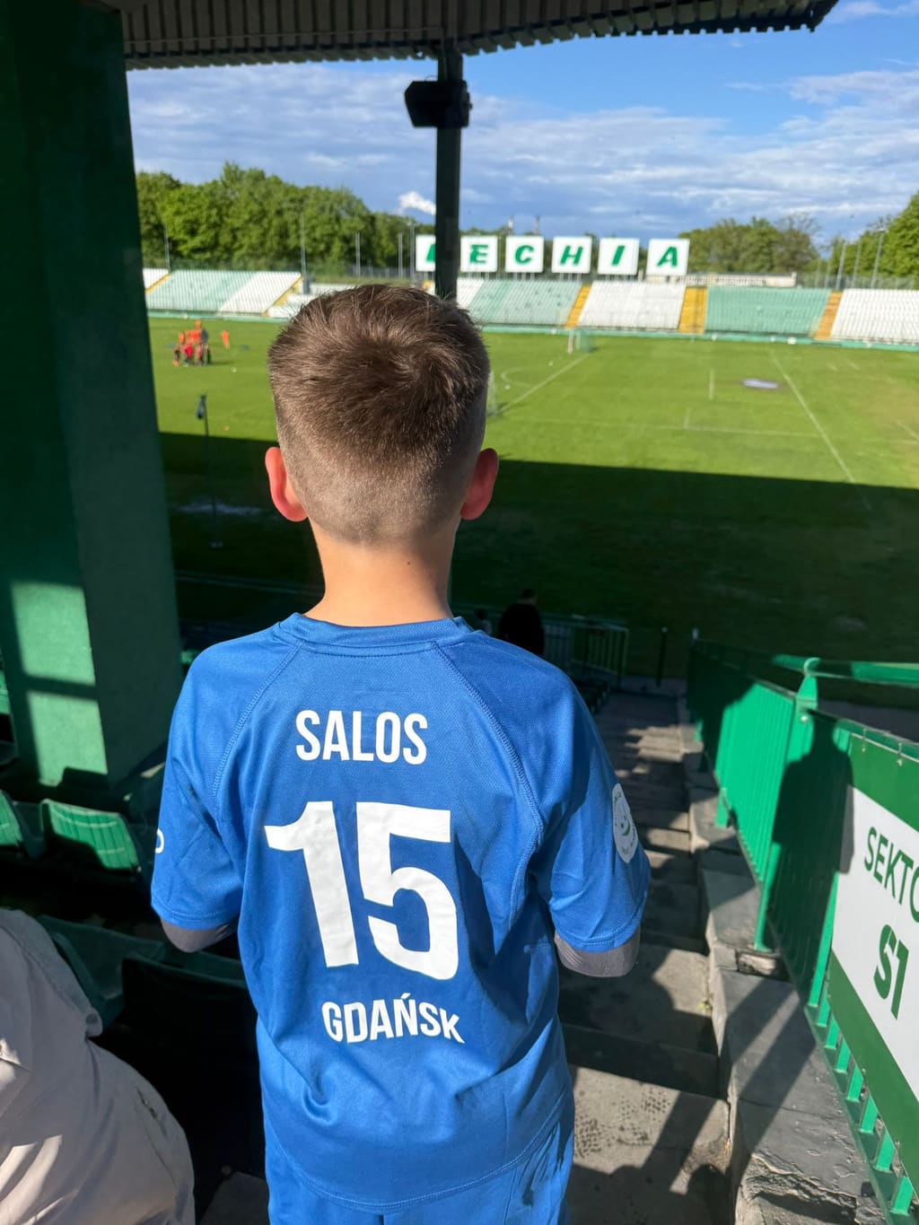 Chłopiec w niebieskiej koszulce SALOS Gdańsk z numerem 15 stoi przy trybunach stadionu Lechii i patrzy na murawę.