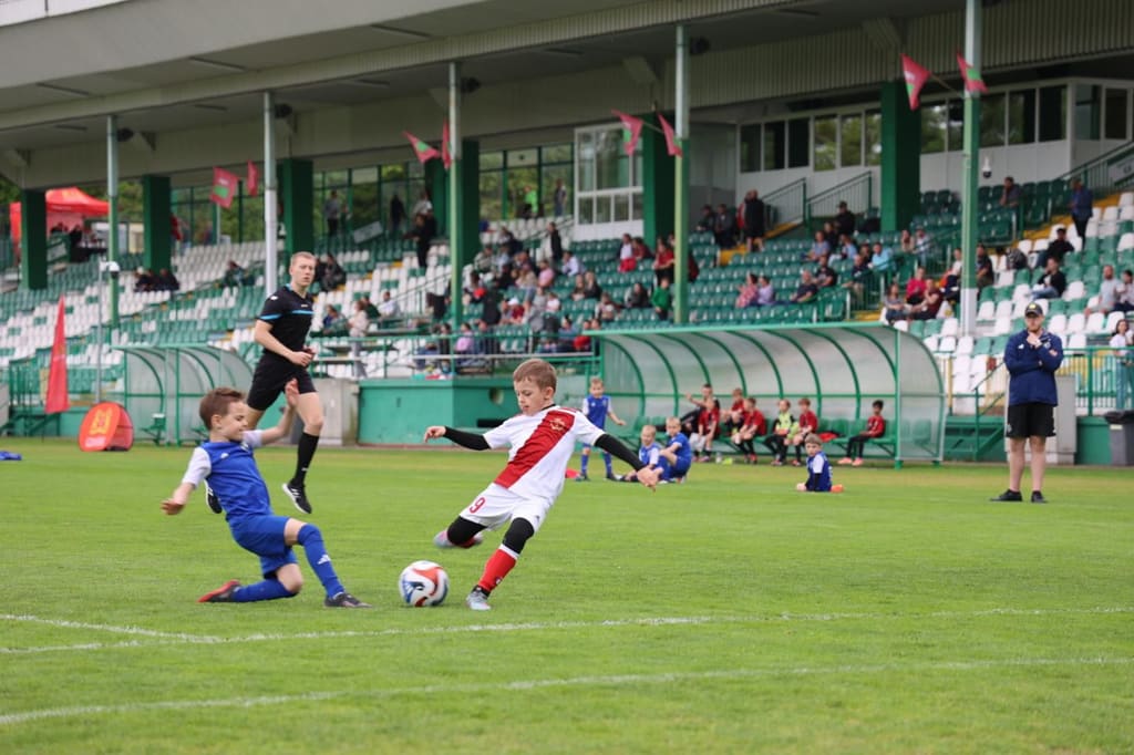 Zawodnik w białym stroju uderza piłkę, obrońca w niebieskim wchodzi w podanie; w tle trybuny stadionu Lechii.