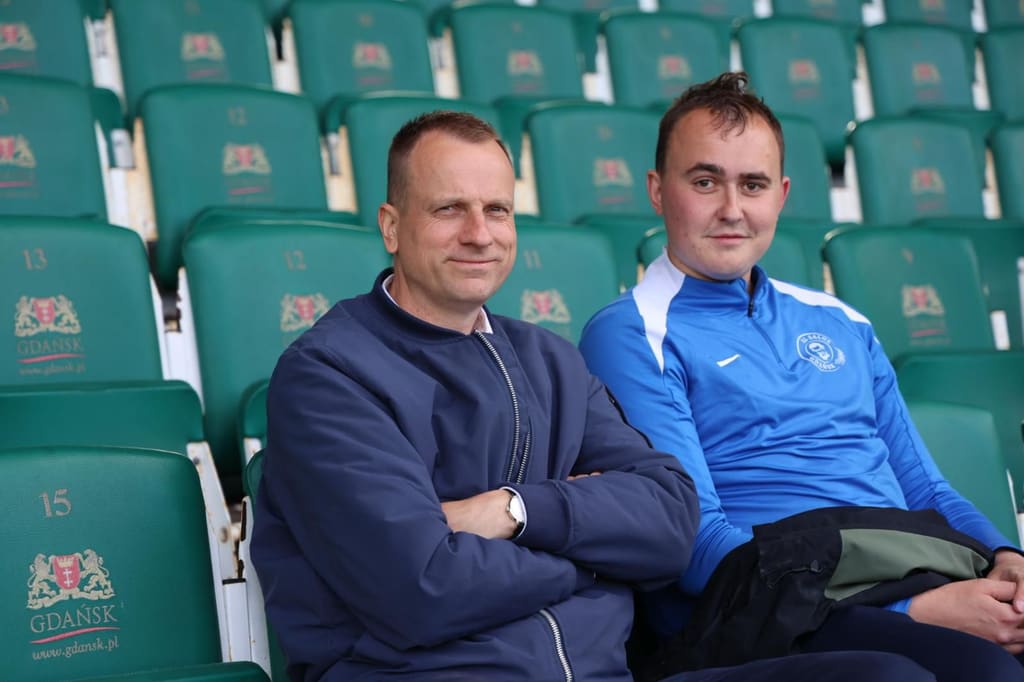 Dwaj trenerzy w klubowych bluzach siedzą na zielonych krzesełkach trybun stadionu Lechii.