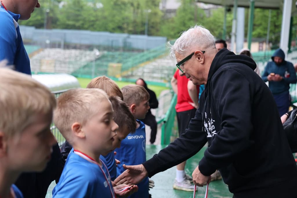 Starszy mężczyzna w czarnej bluzie zakłada medal chłopcu stojącemu w szeregu z innymi zawodnikami.